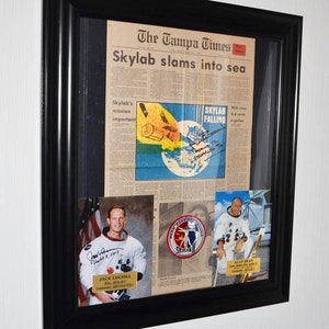May include: A framed newspaper clipping from The Tampa Times with the headline "Skylab slams into sea". The article is about the Skylab space station's re-entry into the Earth's atmosphere. The frame also includes photos of astronauts Jack Lousma and Alan Bean, along with a Skylab II patch.