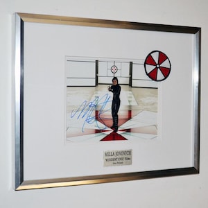 May include: A framed photograph featuring Milla Jovovich in a scene from the Resident Evil films, signed in blue ink. The image includes a red and white circular logo and a plaque below reading Milla Jovovich Resident Evil Films Sony Pictures.
