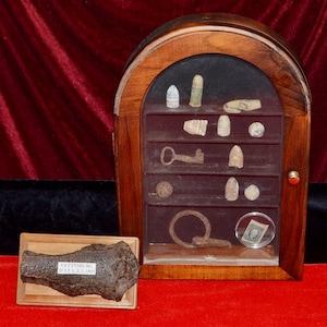 May include: A wooden display case with an arched top containing various metallic artifacts, including bullets, a key, and a ring. Beside it sits a rectangular wooden plaque holding a dark, textured fragment labeled Gettysburg July 1, 2, 3, 1863.