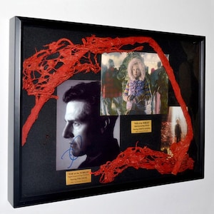 May include: Framed movie memorabilia from "War of the Worlds" featuring Tom Cruise. The display includes photos, signatures, and a vibrant red, textured artistic element. The black frame adds a classic touch to this unique collectible.