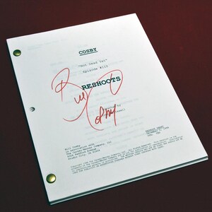 May include: A white script page with the title "COSBY" and the episode title "Not Dead Yet" written in black text. The page is marked "RESHOOTS" in red ink with a red circle around the word. The page also includes a handwritten signature in red ink.