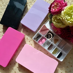 Puede incluir: Cuatro contenedores rectangulares de plástico con tapas en negro, lavanda, rosa y rosa claro. Los contenedores están llenos de varios accesorios para el cabello, incluidos pasadores, cintas para el cabello y pinzas. Los contenedores están dispuestos sobre una superficie con estampado dorado y blanco.