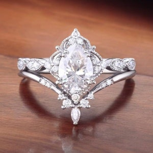 Puede incluir: Un anillo de compromiso de diamante de forma pera con un engaste delicado y ornamentado. El anillo presenta una banda con un dise&ntilde;o en forma de V y un halo de diamantes m&aacute;s peque&ntilde;os que rodean la piedra central.