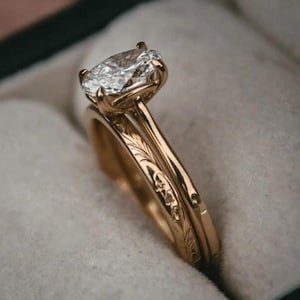 Könnte beinhalten: Ein Paar goldene Eheringe mit einem großen ovalen Diamanten in der Mitte des Verlobungsringes. Die Ringe befinden sich in einer schwarzen Samt-Ringbox.
