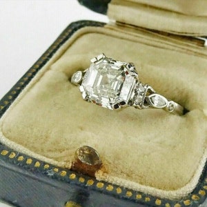 May include: A vintage engagement ring with a large, clear, square-shaped diamond set in a platinum band. The diamond is surrounded by smaller diamonds and the band is decorated with intricate details.