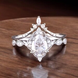 Puede incluir: Un anillo de compromiso de diamante de forma pera con una banda delicada e intrincada. El anillo presenta un engaste en forma de corona con diamantes m&aacute;s peque&ntilde;os que rodean la piedra central.