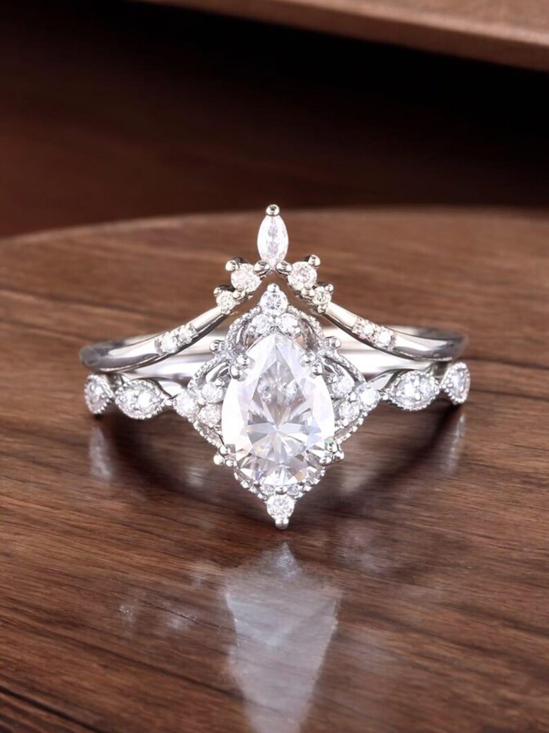 Puede incluir: Un anillo de compromiso de diamante de forma pera con una banda delicada e intrincada. El anillo presenta un engaste en forma de corona con diamantes m&aacute;s peque&ntilde;os que rodean la piedra central.