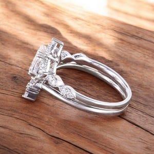Puede incluir: Un anillo de compromiso de plata con una piedra central grande y redonda y piedras m&aacute;s peque&ntilde;as en la banda. El anillo est&aacute; sobre una superficie de madera.