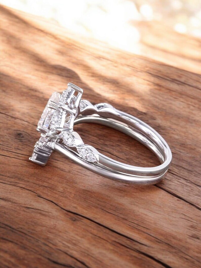 Puede incluir: Un anillo de compromiso de plata con una piedra central grande y redonda y piedras m&aacute;s peque&ntilde;as en la banda. El anillo est&aacute; sobre una superficie de madera.