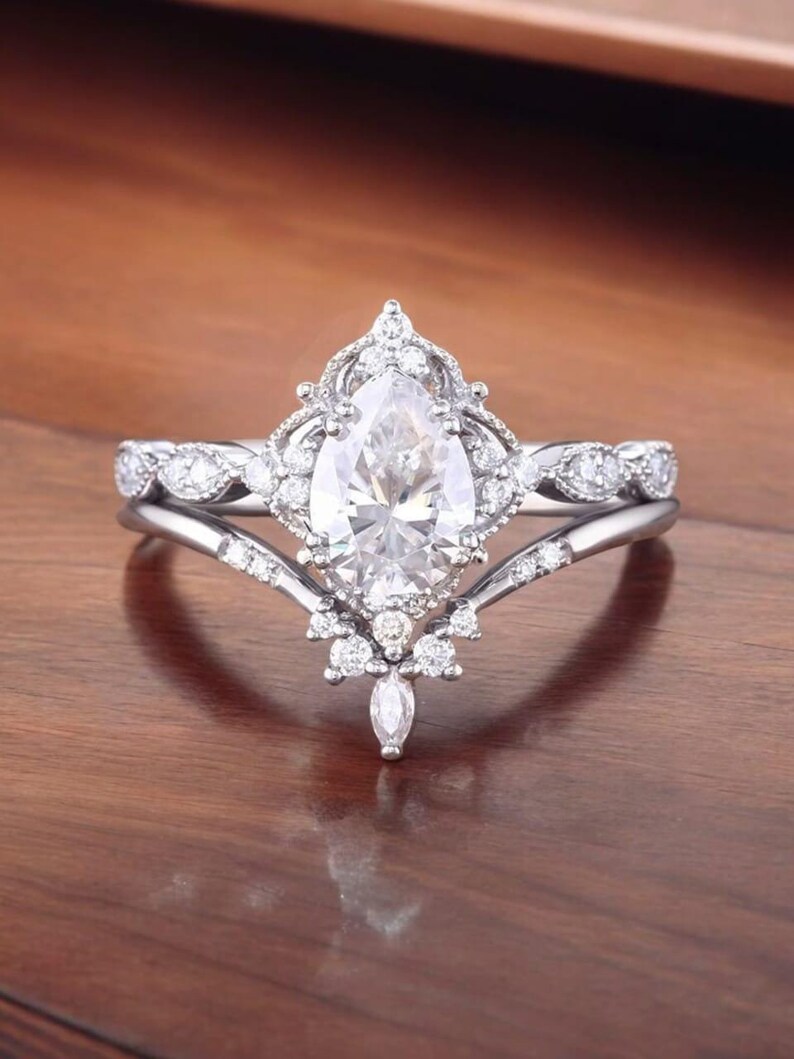 Puede incluir: Un anillo de compromiso de diamante de forma pera con un engaste delicado y ornamentado. El anillo presenta una banda con un dise&ntilde;o en forma de V y un halo de diamantes m&aacute;s peque&ntilde;os que rodean la piedra central.