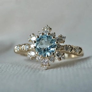 May include: A gold ring with a large, round, light blue aquamarine gemstone surrounded by a cluster of smaller, round, white diamonds. The ring has a delicate, intricate design.