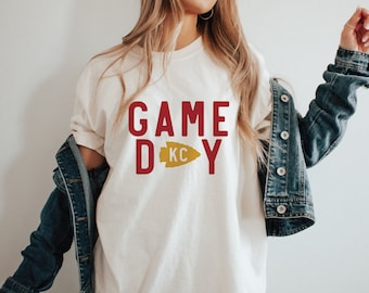 May include: White t-shirt with a red "GAME DAY" graphic and a gold arrow with "KC" inside. The shirt is being worn by a person with long blonde hair and a denim jacket.