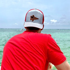May include: A person wearing a red t-shirt and a gray and red baseball cap with an American flag and a jet fighter design. The person is looking out at the ocean.