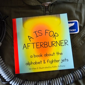 May include: A children's book titled "A Is For Afterburner" about the alphabet and fighter jets. The book has a colorful cover with the title in large letters. The book is on a green flight suit.