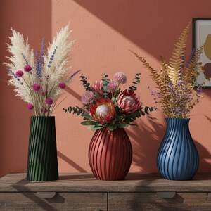May include: Three decorative vases with floral arrangements on a wooden surface. The vases are green, red, and blue. The flowers include pampas grass, allium, protea, and golden ferns. A framed abstract art piece hangs on the wall.