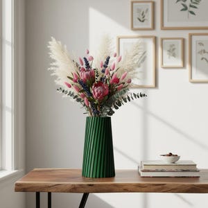 May include: A vibrant bouquet of dried flowers in a green, ribbed vase. The arrangement includes pampas grass, pink protea, and other colorful blooms. The vase sits on a wooden table, with framed botanical prints in the background.