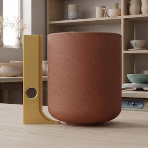 May include: A cylindrical terracotta-colored ceramic vessel with a textured surface, positioned next to a vertical yellow support structure featuring a circular cutout, resting on a light-colored wooden surface in a studio setting.