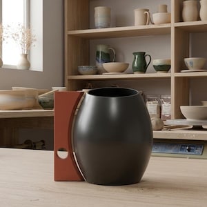 Peut inclure: Une carafe en céramique noir mat avec un corps arrondi et bombé, dotée d'une poignée plate de couleur terre cuite avec une découpe circulaire près de la base. Le récipient présente une large ouverture et repose sur une surface en bois clair.
