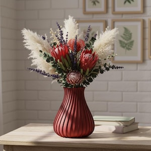 May include: A striking floral display in a red vase. The bouquet includes large red protea flowers, fluffy white pampas grass, and purple lavender. The vase is a deep red with vertical ridges, placed on a light wood table.