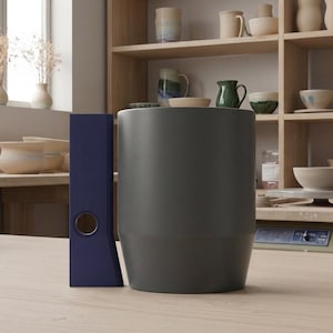 May include: A dark gray cylindrical ceramic vessel with a smooth matte finish positioned next to a tall, narrow, dark blue rectangular object featuring a single circular cutout near the base, both resting on a light-colored wooden surface.