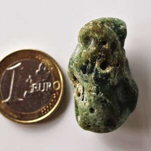 Peut inclure: Une pierre verte rugueuse avec une surface texturée et plusieurs trous. À côté de la pierre se trouve une pièce d'euro, servant de référence de taille. La pierre est de couleur vert clair à foncé.