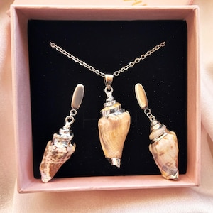 May include: A jewelry set featuring a necklace and earrings, presented in a pink box. The set includes a silver chain necklace with a seashell pendant and matching seashell earrings. The seashells have a natural beige and brown color.