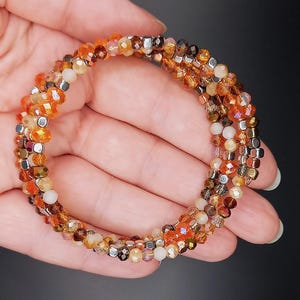 Könnte beinhalten: Ein mehrreihiges Armband aus kleinen, facettierten Glasperlen in Orange-, Braun- und Weißtönen. Die Perlen sind mit silberfarbenen Metallperlen durchsetzt.