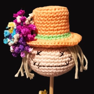 May include: A crocheted scarecrow doll with a pink face, black eyes, and a stitched smile. The doll is wearing a peach-colored hat with a green band and a colorful flower crown.