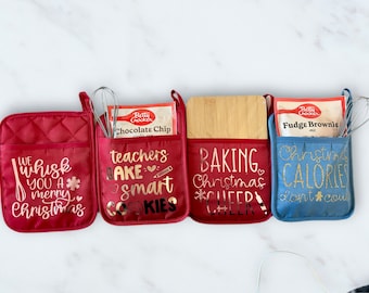 Op de afbeelding: Vier rode en blauwe ovenwantjes met zakken. De wantjes hebben verschillende kerstspreuken in gouden letters. De zakken bevatten bakbenodigdheden, waaronder een doos met chocolade-chip-koekjesmix, een houten snijplank, een doos met fudge-brownie-mix en een garde.