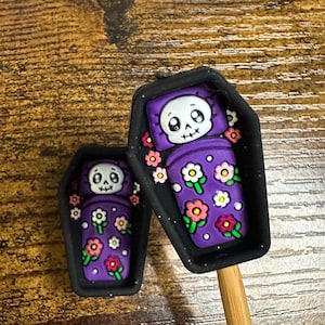 May include: Two black coffin-shaped charms featuring a cartoon skull resting in a purple sleeping bag decorated with colorful flowers. The larger charm is attached to a wooden stick, while the smaller charm is positioned to its left.