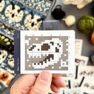 May include: A card with a pixelated Dino T-Rex Skull design, held by a hand. The card has a grid pattern with numbers and the text "24 sts" and "Dino T-Rex Skull". Various balls of yarn and knitting patterns are in the background.