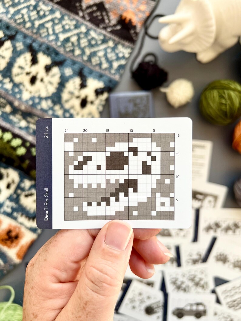 May include: A card with a pixelated Dino T-Rex Skull design, held by a hand. The card has a grid pattern with numbers and the text "24 sts" and "Dino T-Rex Skull". Various balls of yarn and knitting patterns are in the background.