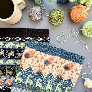 May include: Two knitted cowls with colorful geometric patterns, alongside balls of yarn in various colors, including green, orange, and gray. A ceramic mug and plush dinosaur toys are also present, suggesting a cozy, craft-themed setting.
