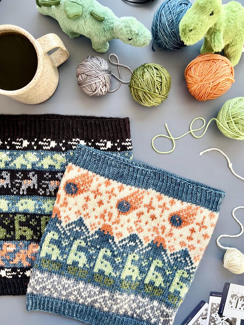 May include: Two knitted cowls with colorful geometric patterns, alongside balls of yarn in various colors, including green, orange, and gray. A ceramic mug and plush dinosaur toys are also present, suggesting a cozy, craft-themed setting.
