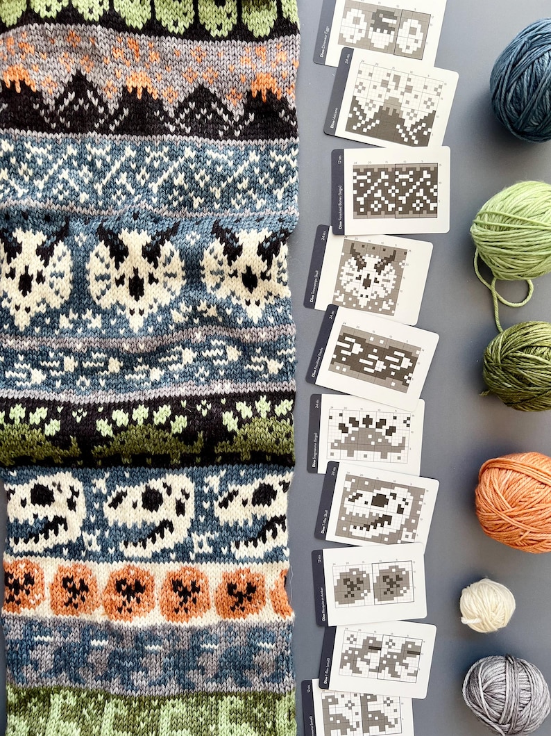 May include: A knitted scarf with a detailed pattern featuring owls, pumpkins, and mountain designs in shades of green, blue, orange, and black. Accompanying the scarf are several pattern cards and balls of yarn.