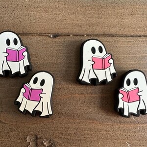 May include: Four white ghost-shaped charms with black outlines and eyes, each holding a pink book. The charms are arranged on a wooden surface. These Halloween-themed charms are perfect for adding a spooky touch to accessories.