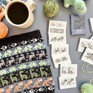 May include: A cozy scene featuring a black, blue, and orange knitted cowl with a dinosaur pattern, a mug of coffee, yarn balls, and a plush dinosaur toy. The image also includes a "Dino Doodle Card Deck" and knitting pattern cards.