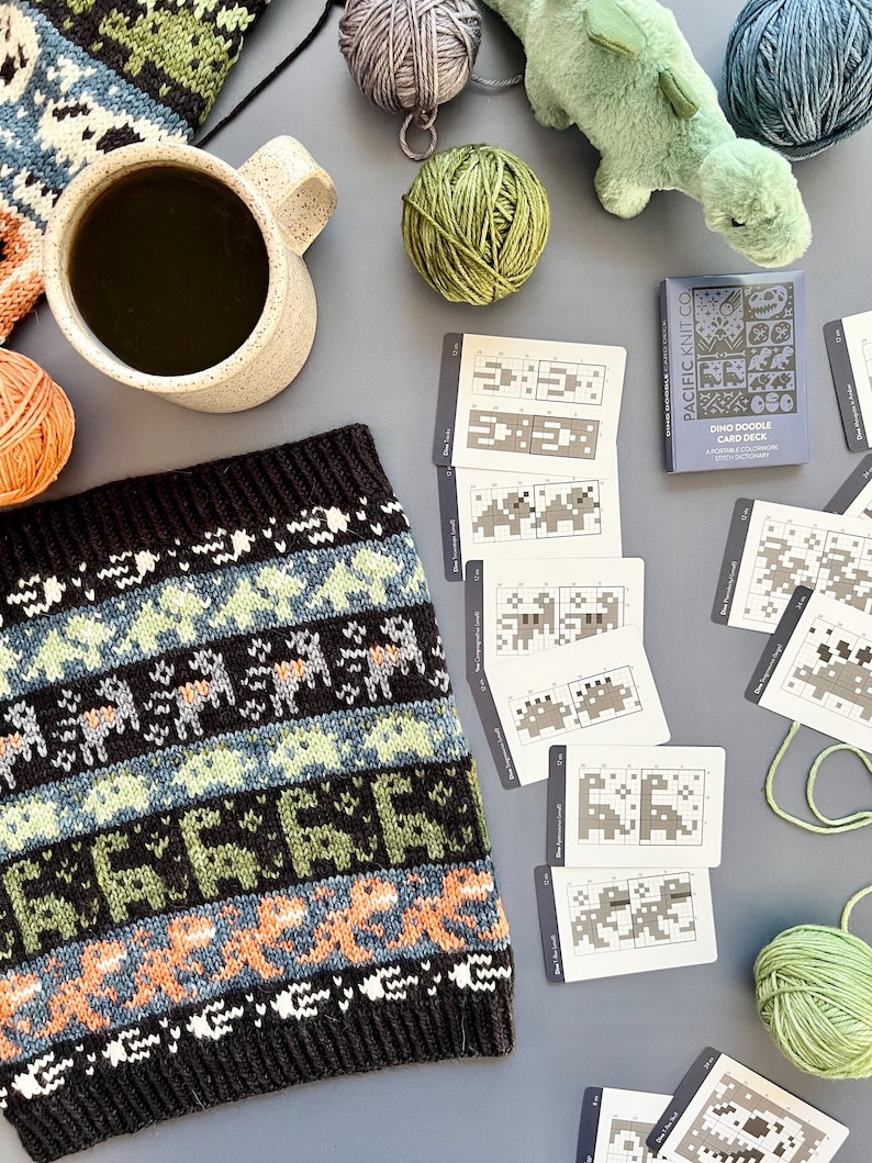 May include: A cozy scene featuring a black, blue, and orange knitted cowl with a dinosaur pattern, a mug of coffee, yarn balls, and a plush dinosaur toy. The image also includes a "Dino Doodle Card Deck" and knitting pattern cards.