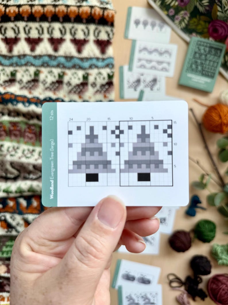 Peut inclure: Une main tient une carte blanche avec un graphique d'arbre de No&euml;l pix&eacute;lis&eacute; intitul&eacute; "Woodland Evergreen Tree (large)". Le graphique est en noir, gris et blanc. D'autres cartes et de la laine de diff&eacute;rentes couleurs sont en arri&egrave;re-plan.