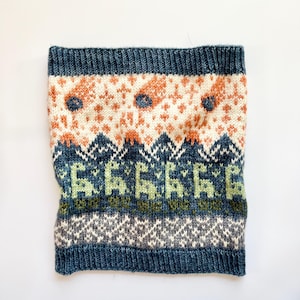 May include: A knitted neck warmer in shades of blue, cream, orange, and green. The design features geometric patterns and stylized floral motifs. The top and bottom edges are a solid dark blue.