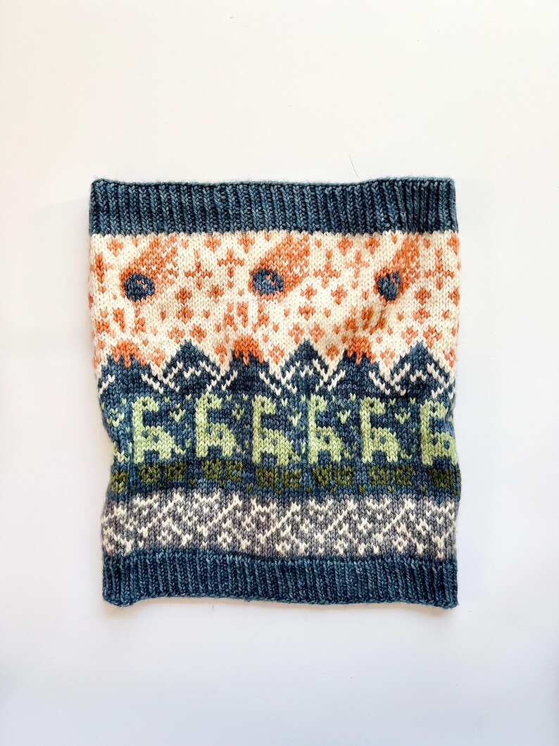 May include: A knitted neck warmer in shades of blue, cream, orange, and green. The design features geometric patterns and stylized floral motifs. The top and bottom edges are a solid dark blue.