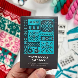 May include: A hand holds a black card deck from Pacific Knit Co. with teal snowflake and tree designs. The text reads "Winter Doodle Card Deck: A Portable Colorwork Stitch Dictionary." Knitting supplies, including yarn and scissors, are in the background.