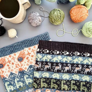 May include: A still-life image featuring two knitted cowls with geometric patterns in blue, orange, and green. Yarn balls in various colors, a mug of coffee, and plush dinosaur toys are scattered around the cowls, suggesting a crafting theme.