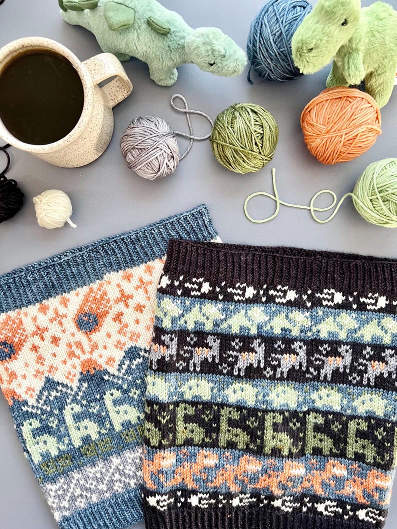 May include: A still-life image featuring two knitted cowls with geometric patterns in blue, orange, and green. Yarn balls in various colors, a mug of coffee, and plush dinosaur toys are scattered around the cowls, suggesting a crafting theme.