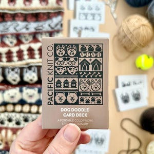 May include: A tan card deck titled "DOG DOODLE CARD DECK" with dog-themed illustrations. The deck is held near a knitted dog-patterned scarf and yarn balls. The text "PACIFIC KNIT CO." and "A PORTABLE COLORWORK STITCH DICTIONARY" are also visible.