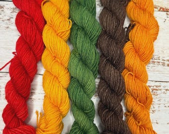 May include: Five skeins of yarn in red, yellow, green, brown, and orange colors. The yarn is arranged in a row on a white wooden surface.