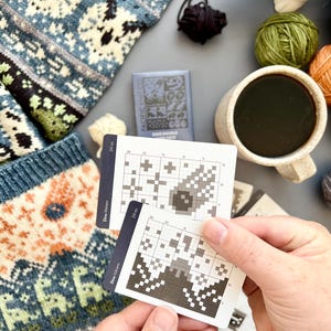 May include: A close-up of knitting supplies, including two cards with pixelated designs, yarn balls in green and orange, and a mug of coffee. A knitted sweater with a blue and white pattern is visible. The cards have the text "Dino Meteor" and "Dino Volcano".