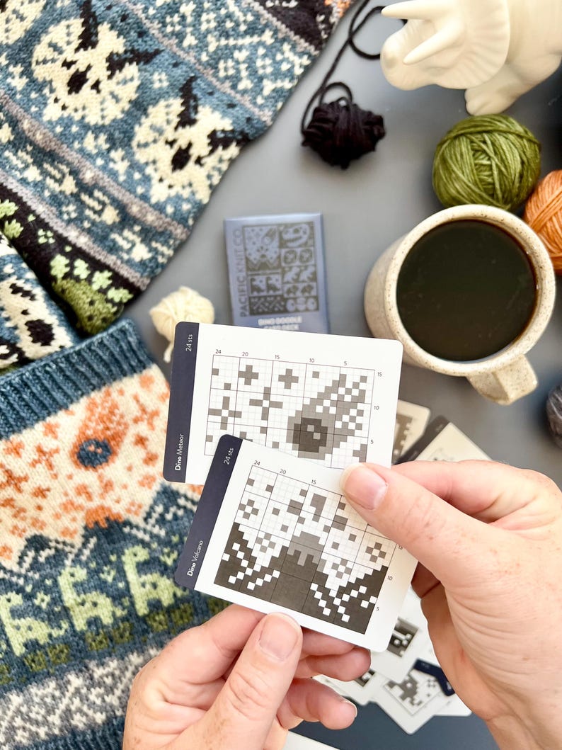 May include: A close-up of knitting supplies, including two cards with pixelated designs, yarn balls in green and orange, and a mug of coffee. A knitted sweater with a blue and white pattern is visible. The cards have the text "Dino Meteor" and "Dino Volcano".