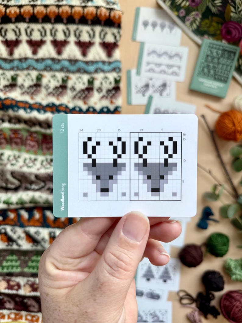 Peut inclure: Une main tient une carte avec un motif de cerf pix&eacute;lis&eacute;, intitul&eacute;e "Woodland Stag". Des fournitures de tricot, dont de la laine et des exemples de motifs, entourent la carte. Un pull tricot&eacute; avec un motif similaire est visible.
