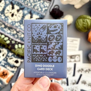 May include: A blue card deck titled "Dino Doodle Card Deck" by Pacific Knit Co. The card deck features dinosaur-themed illustrations. The text on the card reads "A portable colorwork stitch dictionary". The background includes yarn and knitted fabric.